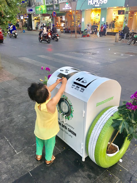 Một em nhỏ được mẹ hướng dẫn bỏ lá cây vào ngăn phân loại rác hữu cơ