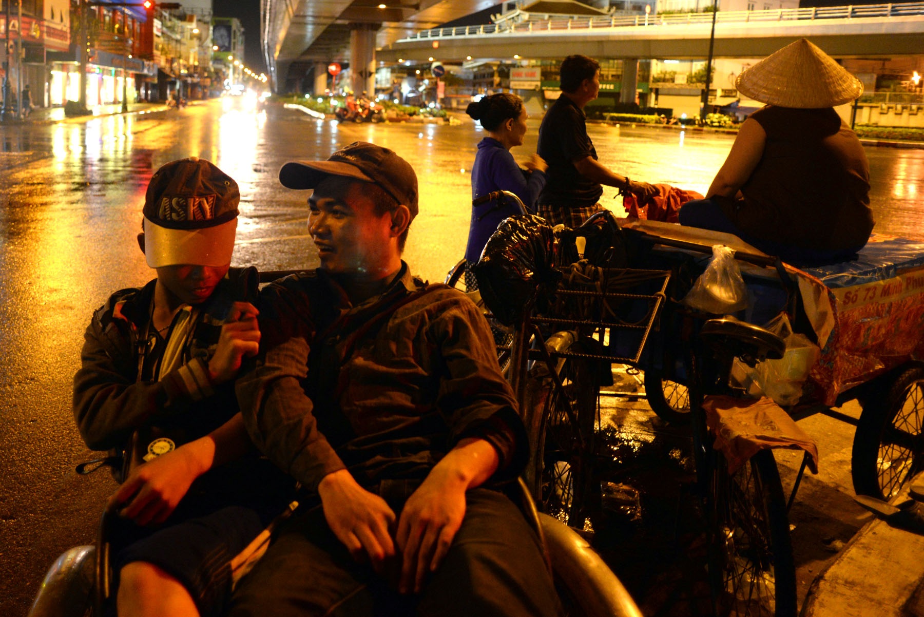 Những phận người nghèo trong đêm mưa Sài Gòn - 3
Hai thanh thiếu niên khác quê nhưng đồng cảnh kiếm sống bên vỉa hè nên thân thiết như anh em. Ngoan (20 tuổi, quê ở Bến Tre) làm nghề đạp xích lô. Huy (14 tuổi, quê Phú Yên) đi bán vé số. Ngoan chia sẻ: “Trời mưa không có khách gọi chở đồ, thằng Huy cũng mới bán xong xấp vé số nên chạy lại chơi với em cho đỡ buồn. Bình thường người ta cho đồ, hai anh em đều để phần cho nhau”.