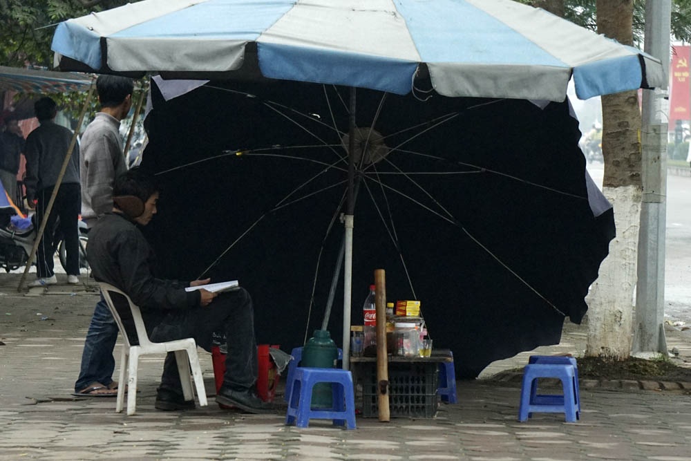 Người Hà Nội co ro trong đợt rét mạnh nhất kể từ đầu mùa - 7 Một quán cóc ven đường nghiêng ô không để che nắng mà để chắn đợt gió mùa ào ạt thổi.