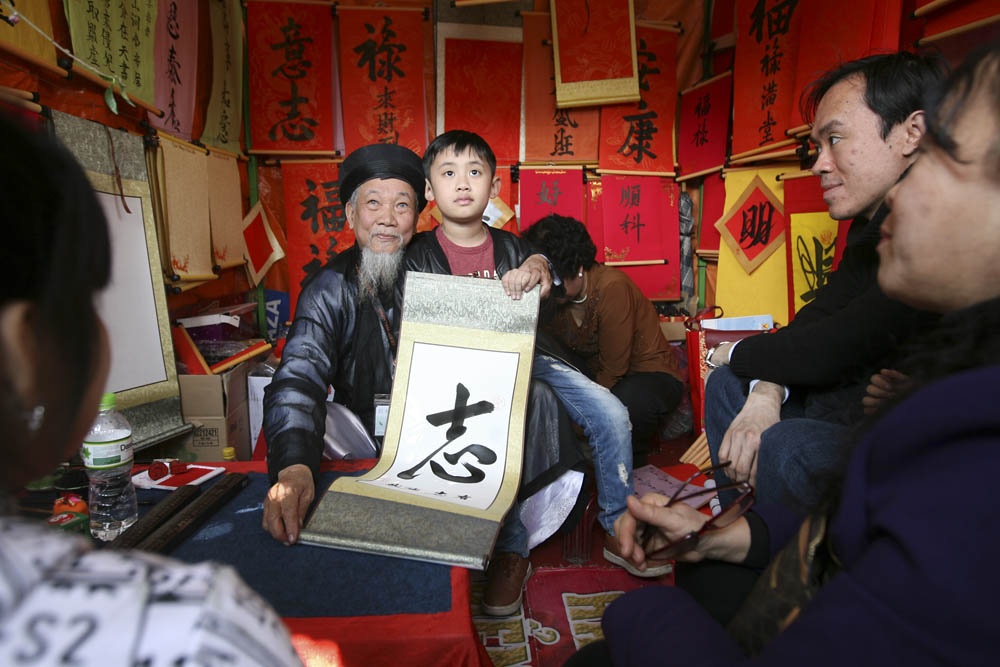 Xin chữ đầu năm, "ông đồ" vắng, "ông đồ "đông" - 3 Ông đồ Nguyễn Mạnh Hùng thuộc CLB thư pháp Hương Nam khá đông khách, khách hàng nhí sau khi có chữ không quên chụp ảnh lưu niệm cùng ông đồ.