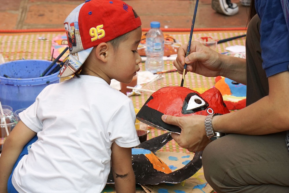 Hàng trăm em nhỏ tập làm mặt nạ giấy bồi chơi Tết Trung thu - 4 Một em nhỏ theo dõi người lớn hướng dẫn cách vẽ mặt nạ.