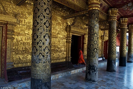 Một chú tiểu bước vào ngôi đền Phật giáo Wat Mai ở Luang Prabang, Lào. Trong ảnh là một lối vào dát vàng được trang trí cầu kỳ, tỉ mỉ, và được bê đỡ bằng các hàng cột lớn.