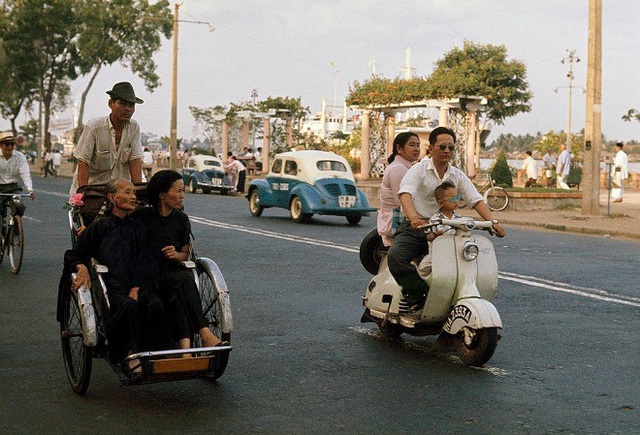 Người đàn ông chở vợ và con trai dạo phố trên chiếc xe Vespa màu trắng bên cạnh hai bà cụ đi xích lô ngắm phố phường. Ảnh chụp ở bến Bạch Đằng năm 1961.