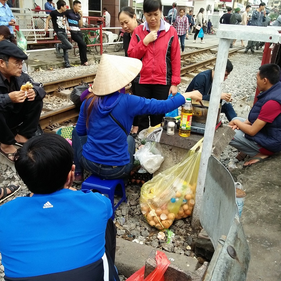 Hà Nội: Hãi hùng cảnh "họp chợ" trên đường ray tàu hỏa - 2 Trong khi người bán bày biện đủ loại dụng cụ, xoog nồi, tủ đựng bánh mỳ, bếp than… trên đường ray thì người mua cũng vô tư ngồi ngay bên cạnh để thưởng thức.