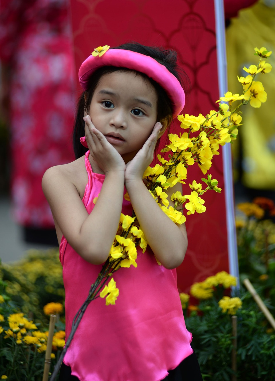 Em bé xinh xắn với trang phục áo yếm, cành hoa mai rực rỡ tạo dáng trước ống kính.