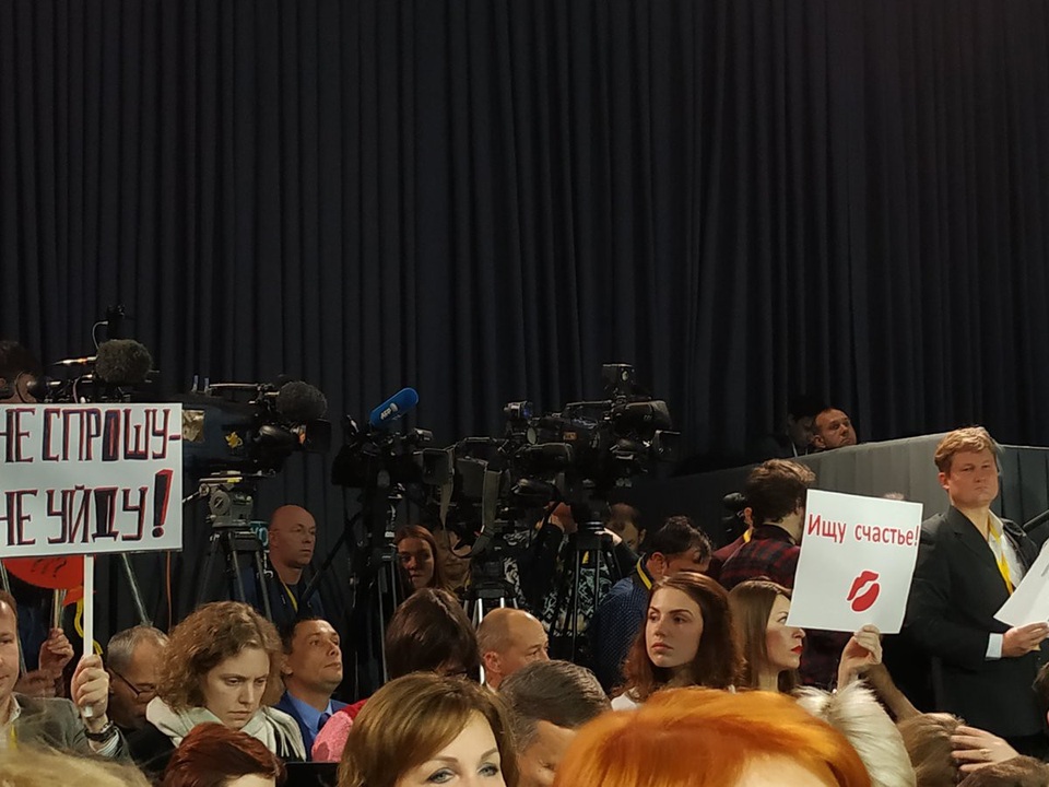 Những bức ảnh đăng trên Twitter cho thấy một nhà báo giơ tấm biển với dòng chữ “đợi chờ hạnh phúc mỉm cười” với hình ảnh một nụ hôn ở dưới. Trong khi đó, người đứng cách cô vài hàng giơ tấm biển thể hiện sự quyết tâm với dòng chữ: “Không hỏi, không về”.