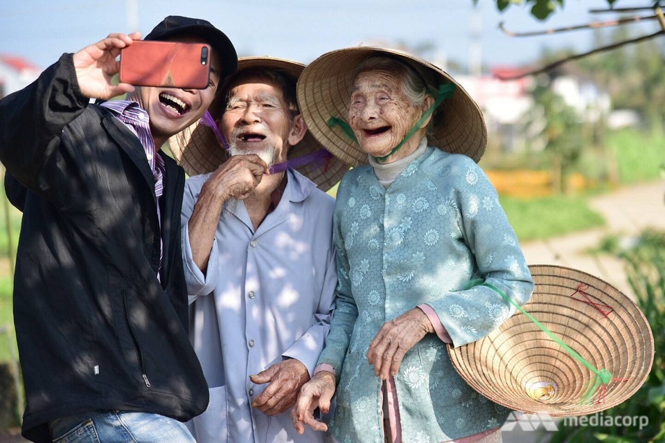 Báo nước ngoài viết về cặp vợ chồng già giữ nghề trồng rau sạch ở Việt Nam - 2 Cụ So và cụ Loi chụp ảnh tự sướng với khách du lịch. (Ảnh: Mediacorp)