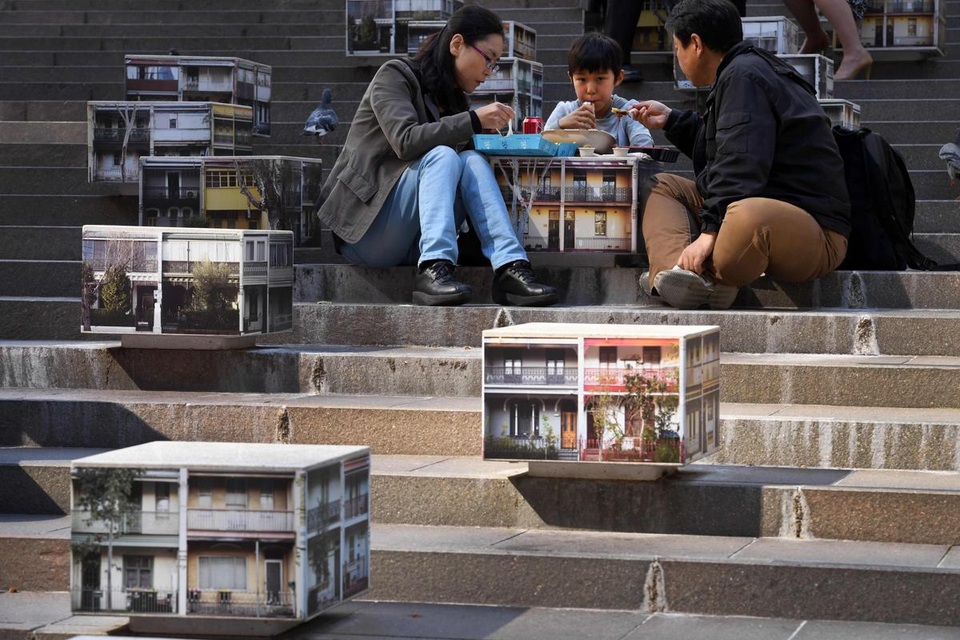 Một gia đình đang ngồi ăn trưa bên một bệ đá được trang trí bằng những bức ảnh chụp lại những ngôi nhà tập thể cũ kỹ. Ảnh chụp tại khu phố dành cho người đi bộ Martin Place ở thành phố Sydney, Úc.